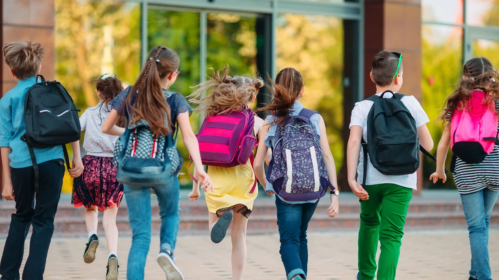 Seven children with backpacks walking and running toward school doors, seen from behind