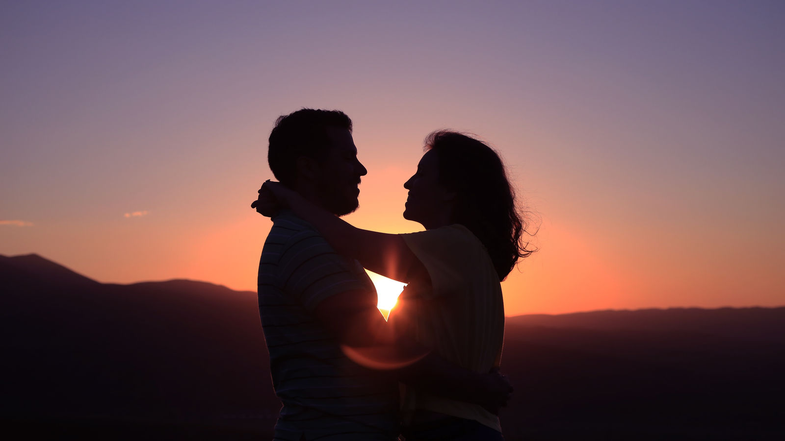 A couple embracing in silhouette against an orange sunset over distant hills